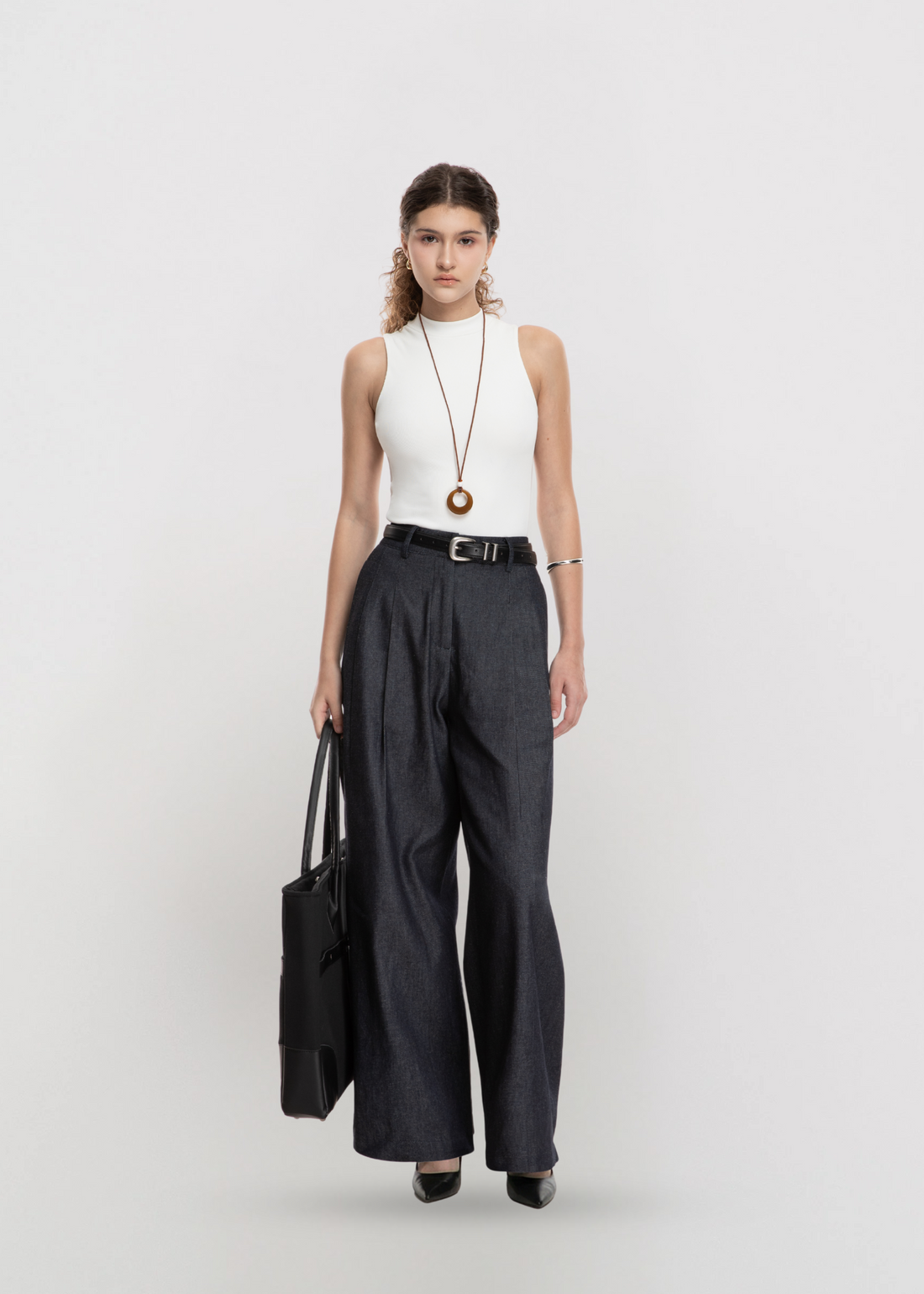 Woman wearing a white sleeveless top and black wide-leg pants, holding a black bag on a light gray background.