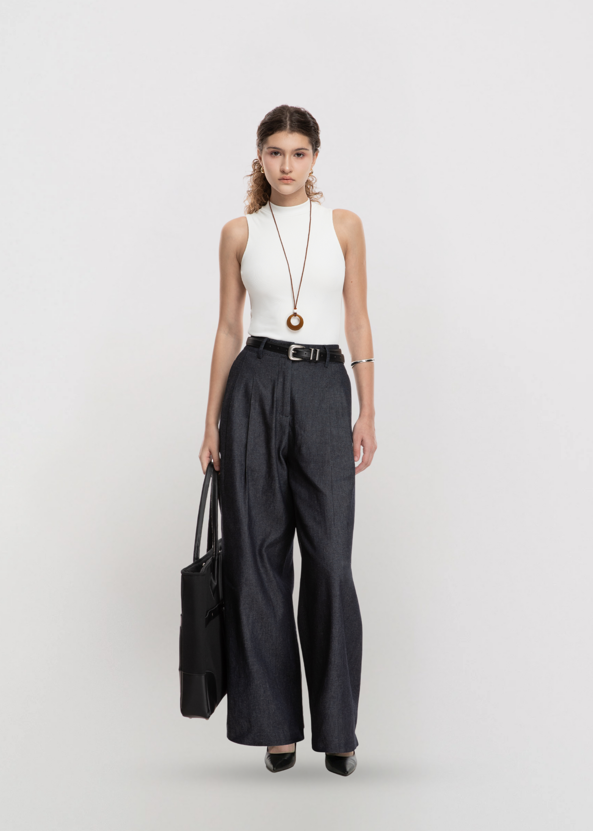 Woman wearing a white sleeveless top and black wide-leg pants, holding a black bag on a light gray background.